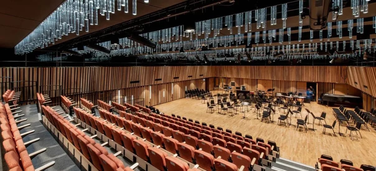 A wide view of a modern concert hall interior, featuring rows of orange-red seats, warm wooden wall paneling, a stage area with chairs and music stands, and a ceiling adorned with numerous glass tube lights.