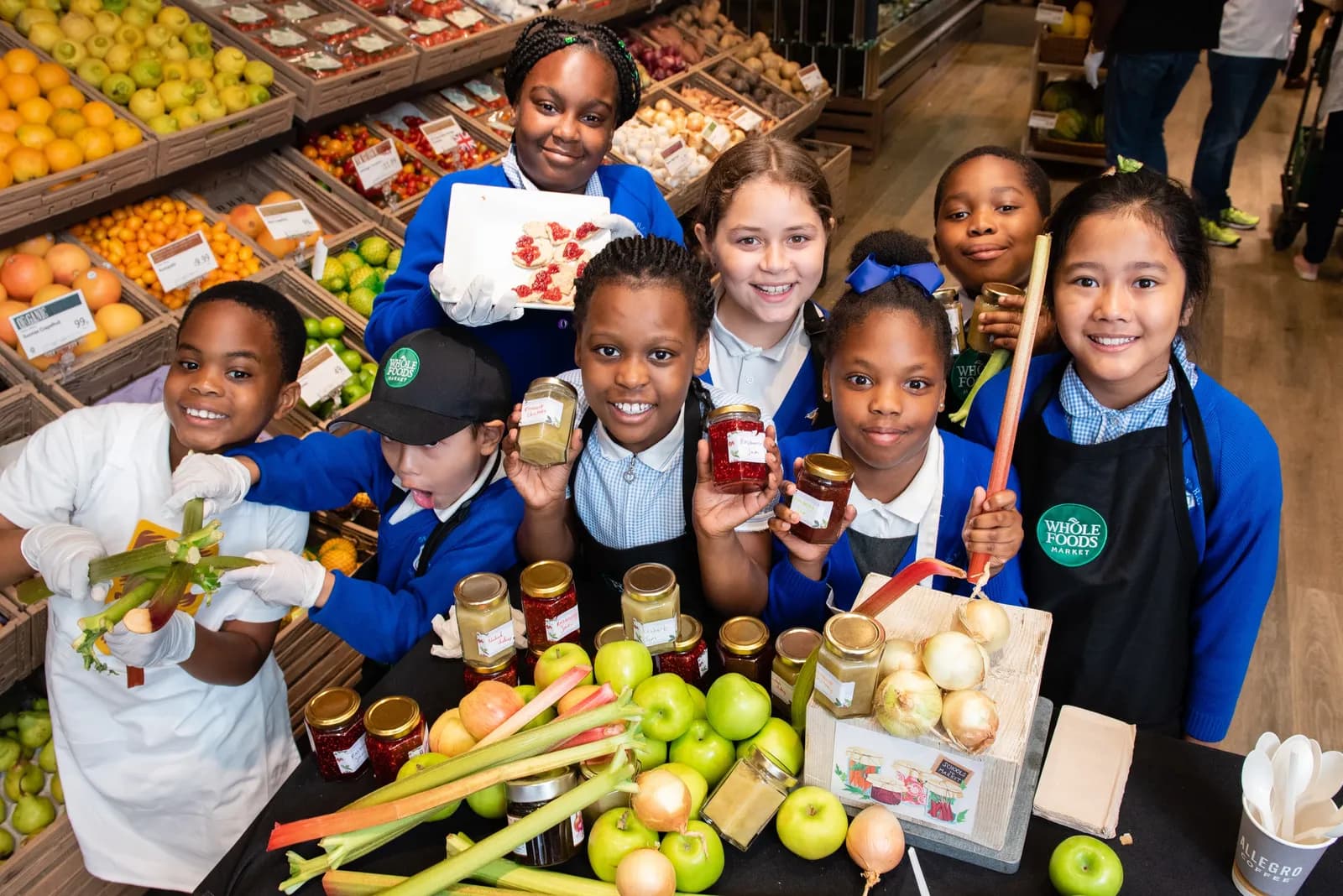 A group of smiling diverse children, dressed in school uniforms and aprons, standing in a Whole Foods Market aisle with produce, holding jars of preserves and fresh produce like apples and rhubarb.